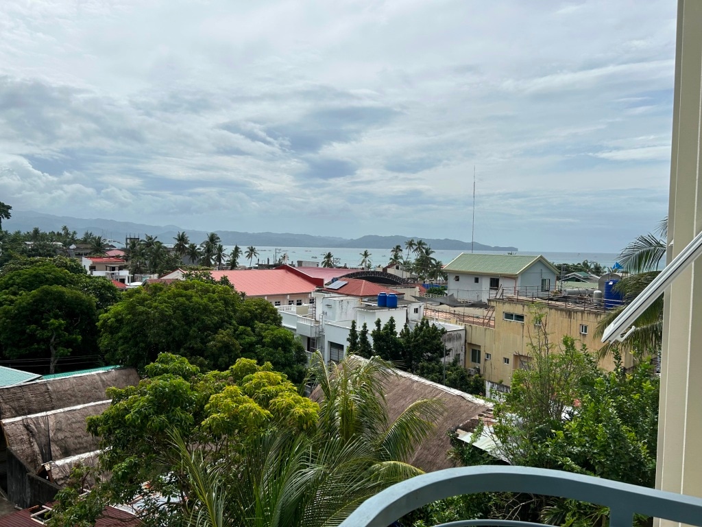 A view from a balcony showcases lush greenery in the foreground with diverse rooftops and distant ocean views. The horizon is lined with hills under a cloudy sky, creating a serene coastal atmosphere.