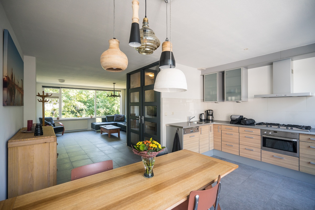 An open kitchen area features modern cabinetry and stainless steel appliances. A wooden dining table is complemented by several chairs. Large windows allow natural light to brighten the space, while a seating area is visible in the background, creating a relaxed ambiance.