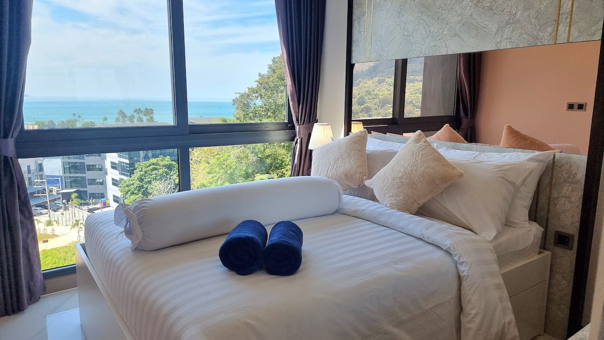 A well-appointed bedroom features a spacious bed with crisp white linens and plush decorative pillows. Large windows offer a panoramic view of the sea and surrounding greenery, allowing natural light to illuminate the room. Two neatly rolled towels are presented on the bed.