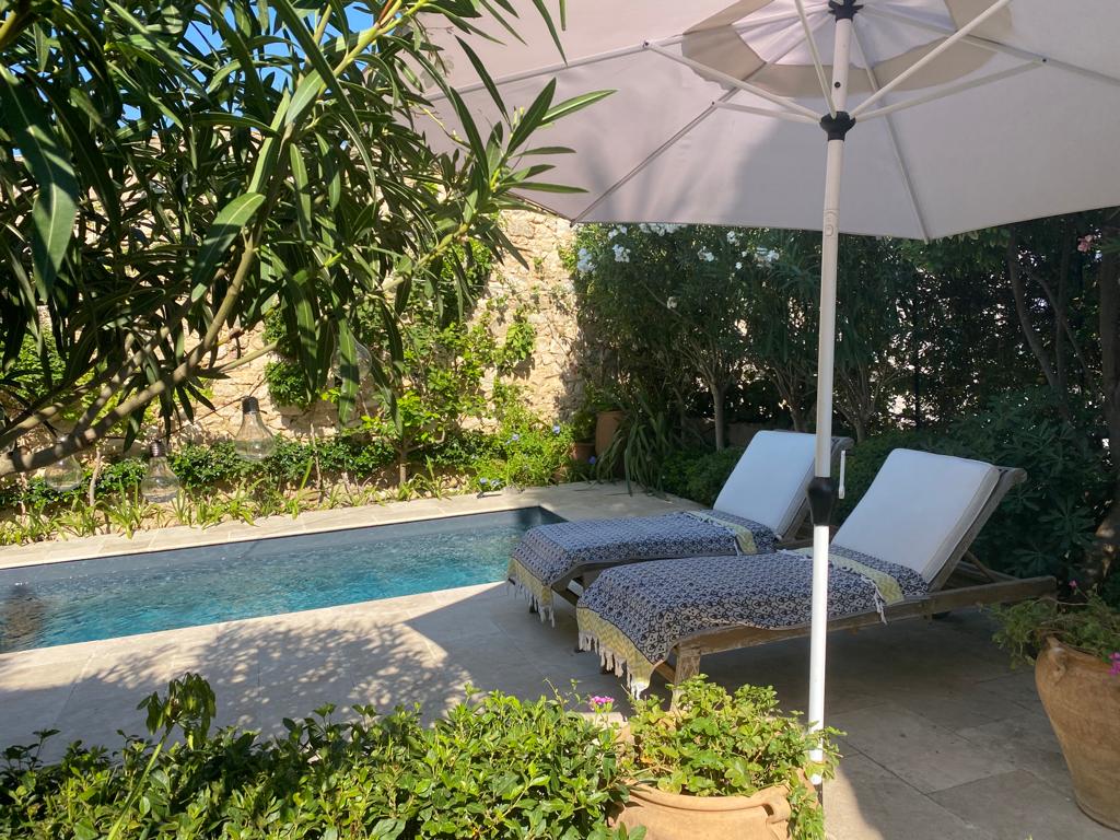 Two sun loungers are positioned under a large umbrella beside a swimming pool. The loungers are draped with patterned textiles, surrounded by lush greenery and flowering plants. Stone walls are visible in the background, enhancing the tranquil poolside setting.