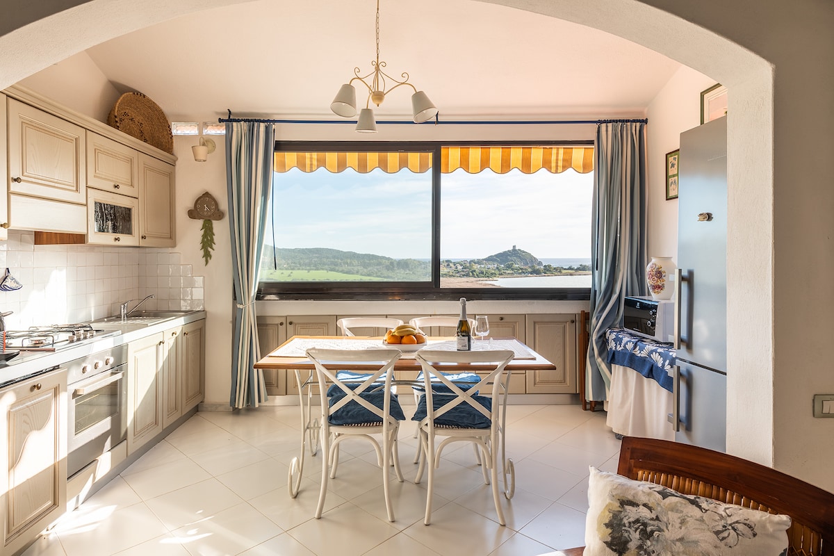 A well-equipped kitchen features a large window with a scenic view of the surrounding landscape. A dining table is set with light-colored chairs, and a fruit bowl and wine bottle are displayed. The space is brightened by natural light and includes modern appliances.