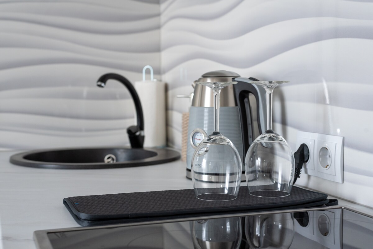 A modern kitchen setup is depicted, featuring a kettle and two inverted wine glasses. The sleek countertop is complemented by a black stovetop and a stylish sink, all set against a wavy textured backsplash.