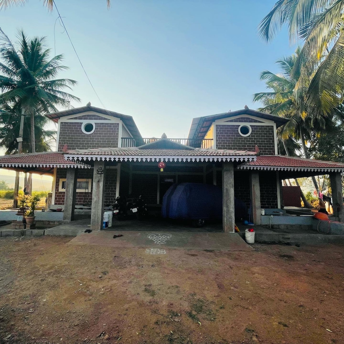 A two-story house is depicted with a tiled roof and unique circular windows. The façade features stone accents and a spacious porch. Surrounding palm trees provide shade, while a gravel pathway leads to the entrance. A vehicle is covered nearby, enhancing the relaxing outdoor setting.