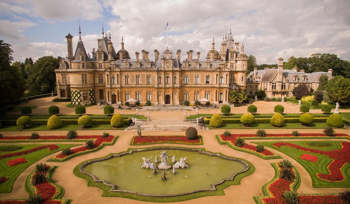 A grand manor house is set amidst meticulously landscaped gardens, featuring vibrant flowerbeds and a large central fountain. The architectural details of the building, including ornate windows and towers, are complemented by the surrounding greenery and neatly trimmed hedges.
