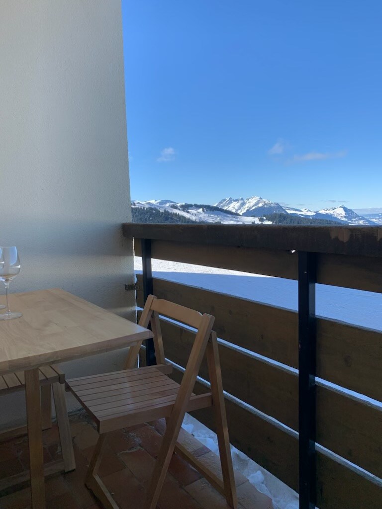 A small balcony features a wooden table and a folding chair, overlooking expansive snow-covered mountains under a clear blue sky. The railing provides an unobstructed view of the scenic landscape, enhancing the peaceful atmosphere.