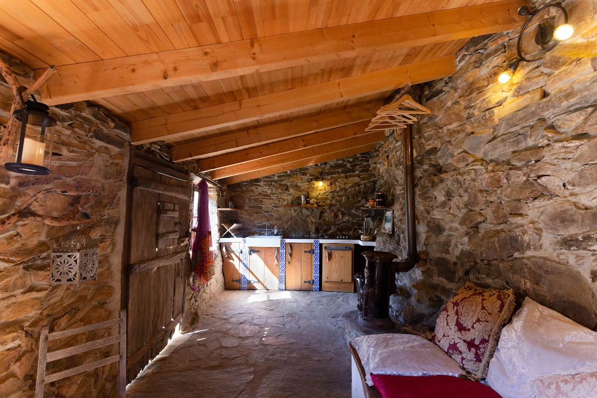 A rustic interior features stone walls complemented by wooden beams and a wooden ceiling. A cozy sitting area is arranged with a throw blanket and pillows on a daybed. A cast-iron wood burner adds warmth, and a kitchenette with wooden cabinets is visible in the background.