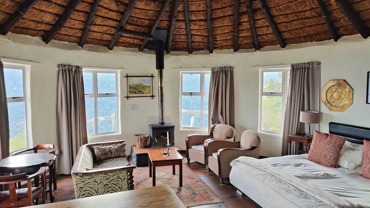 The interior of the round thatched rondavel is characterized by a cozy seating arrangement, including two armchairs, a small table, and a charming couch. Large windows allow natural light and offer views of the surrounding landscape. A decorative rug is positioned on the floor, complemented by a wood-burning stove.