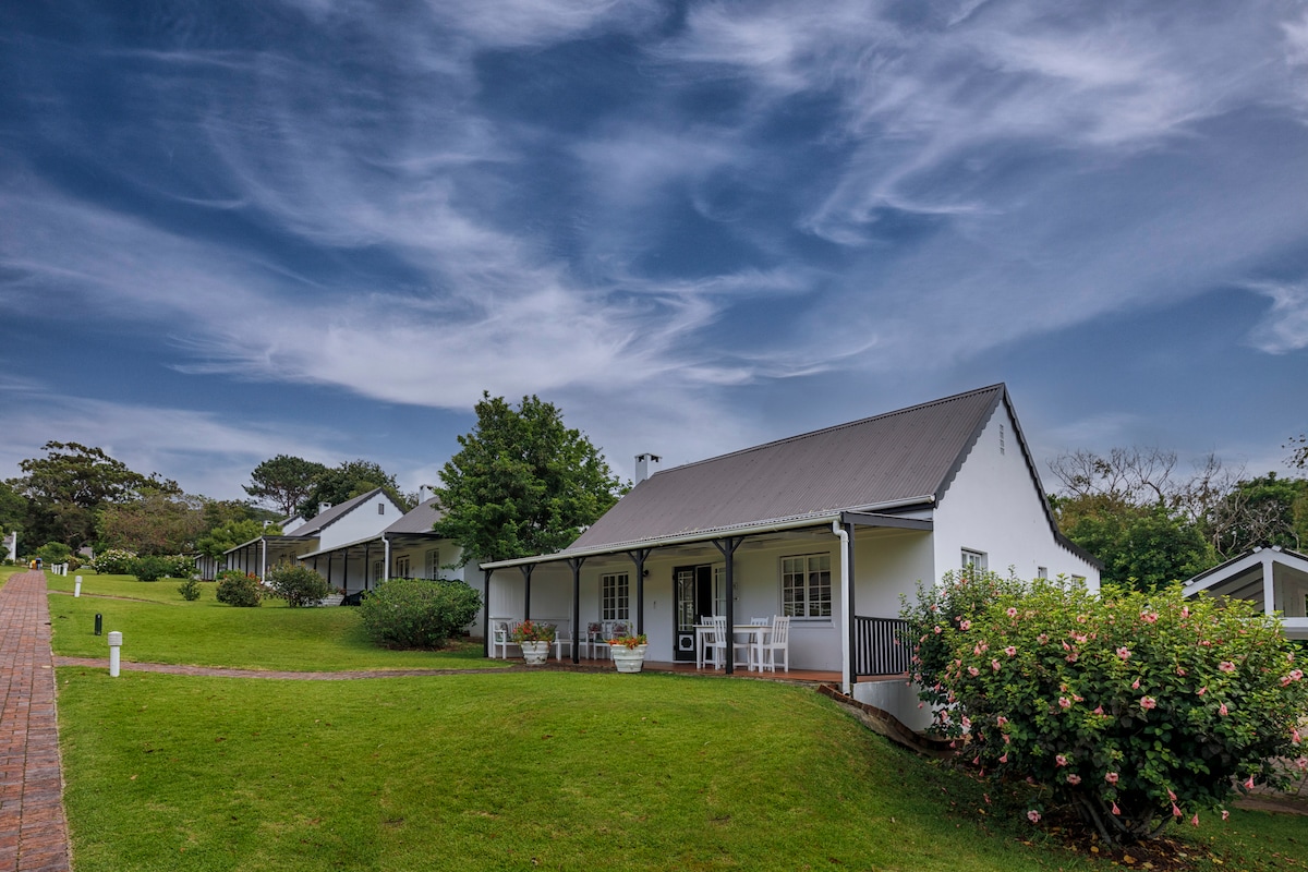 A charming single-storey cottage is set amongst lush greenery, featuring a sloped pathway leading to the entrance. The building has a pitched roof and a welcoming veranda with seating options, surrounded by flowering plants and trees under a sky adorned with wispy clouds.