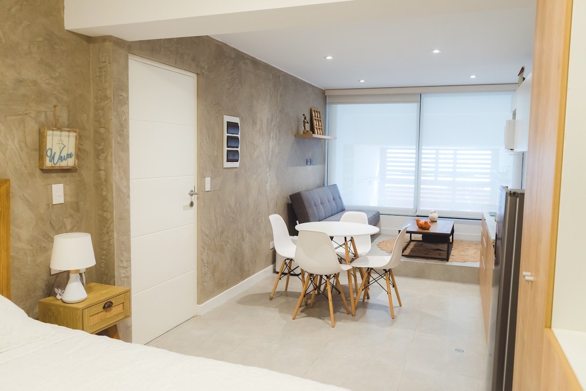 A modern living area showcases a comfortable gray sofa and a light wood dining table surrounded by white chairs. Natural light fills the space through large sliding windows, enhancing the neutral tones of the walls and flooring.