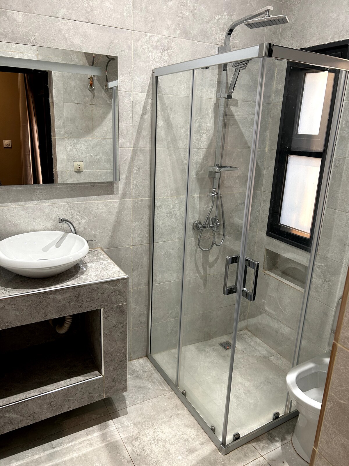 A modern bathroom is shown, featuring a glass shower enclosure with a rainfall showerhead. A sleek countertop holds a round sink, complemented by a wall-mounted mirror. Neutral grey tiles create a cohesive look, while a compact toilet is located next to the enclosed shower.