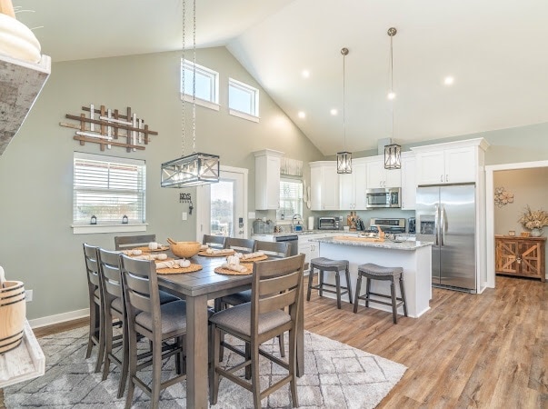 An open-concept kitchen and dining area is highlighted by a large wooden table surrounded by eight chairs. Bright lighting fixtures hang above, and the kitchen features modern appliances and ample counter space. Large windows allow natural light to fill the room.