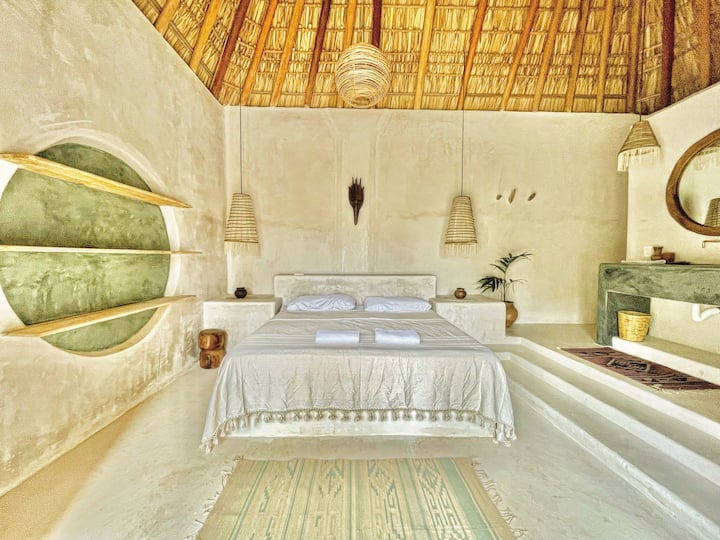 Dhomë komode me tavan të lartë në stil palapa dhe materiale natyrale që krijojnë një atmosferë të ngrohtë dhe çlodhëse. Ajo ka një krevat të madh, ndriçim të butë dhe detaje të punuara me dorë. Ka një banjë private me dush për më shumë rehati.