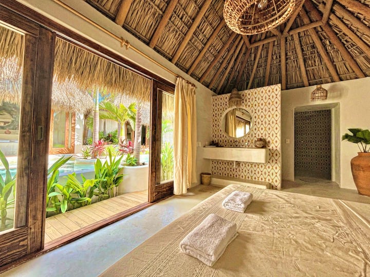 Dhomë në stil tropikal me çati palapa, finiture të punuara me dorë dhe një atmosferë relaksuese ✨ Ka dritare të mëdha me pamje nga zona e jashtme, një banjë private dhe detaje natyrale që krijojnë një hapësirë të ngrohtë dhe mikpritëse