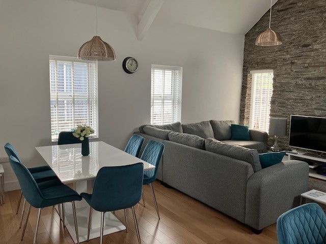 A spacious living and dining area is highlighted by a large grey sofa and a sleek dining table with blue chairs. Natural light filters through the two white-framed windows, while a modern clock and pendant lights add design elements to the room.