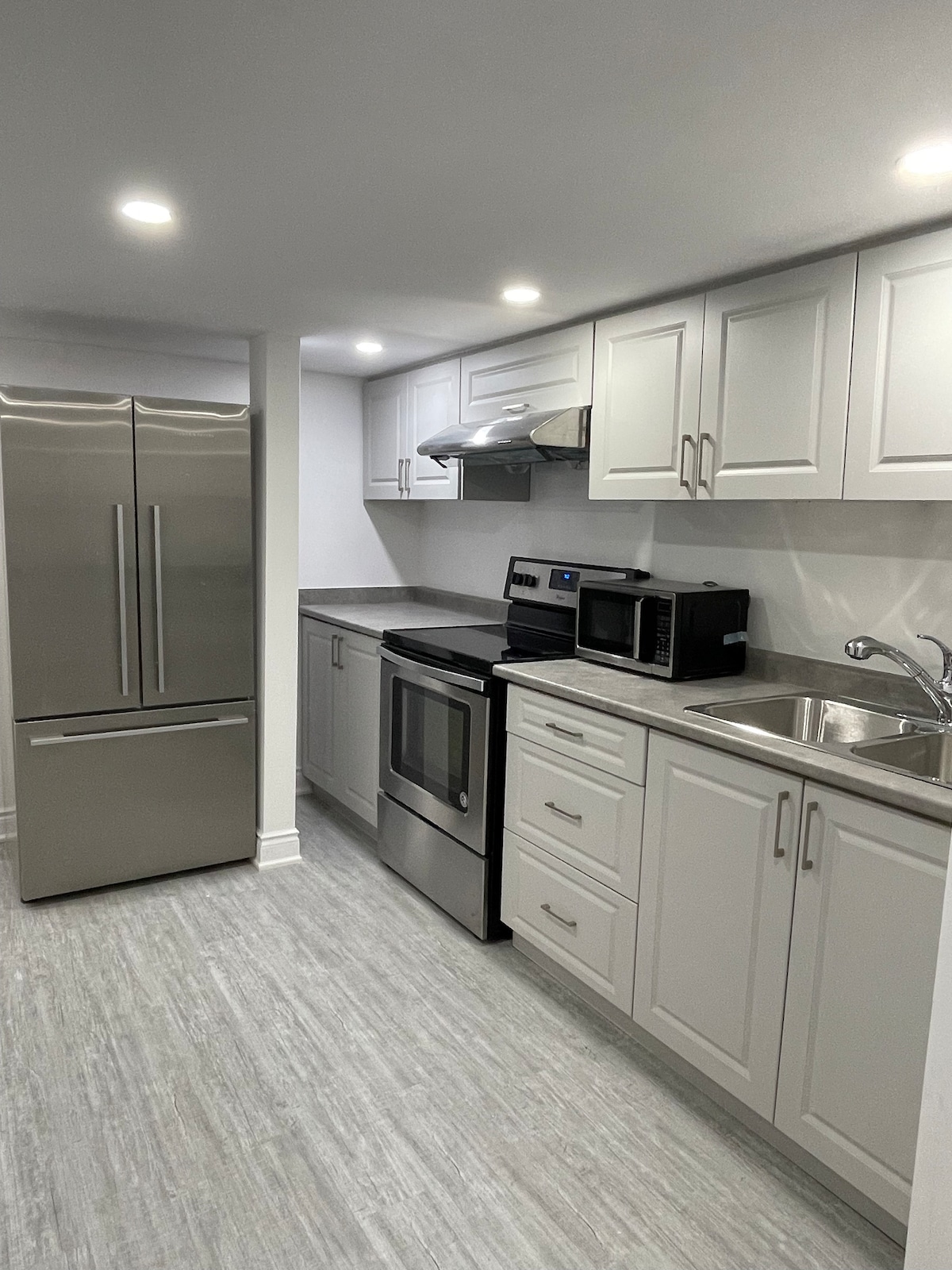 A modern kitchen features stainless steel appliances, including a large refrigerator and a smooth-top stove. White cabinetry and a gray countertop provide a clean and bright cooking space, with a microwave and sink neatly arranged for convenience.