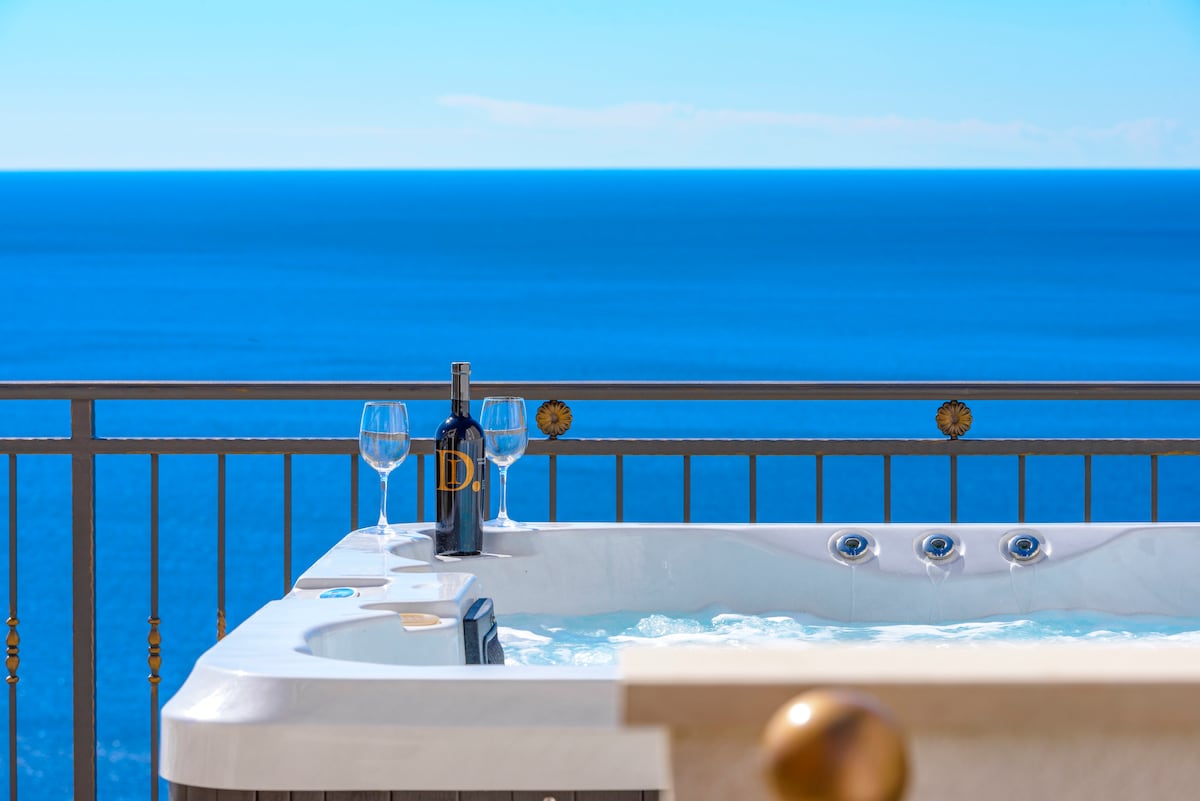 A hot tub with jets is positioned on a balcony overlooking the expansive ocean. Accompanied by a bottle of wine and two glasses, the scene emphasizes relaxation with the clear blue water in the background, creating a serene getaway atmosphere.