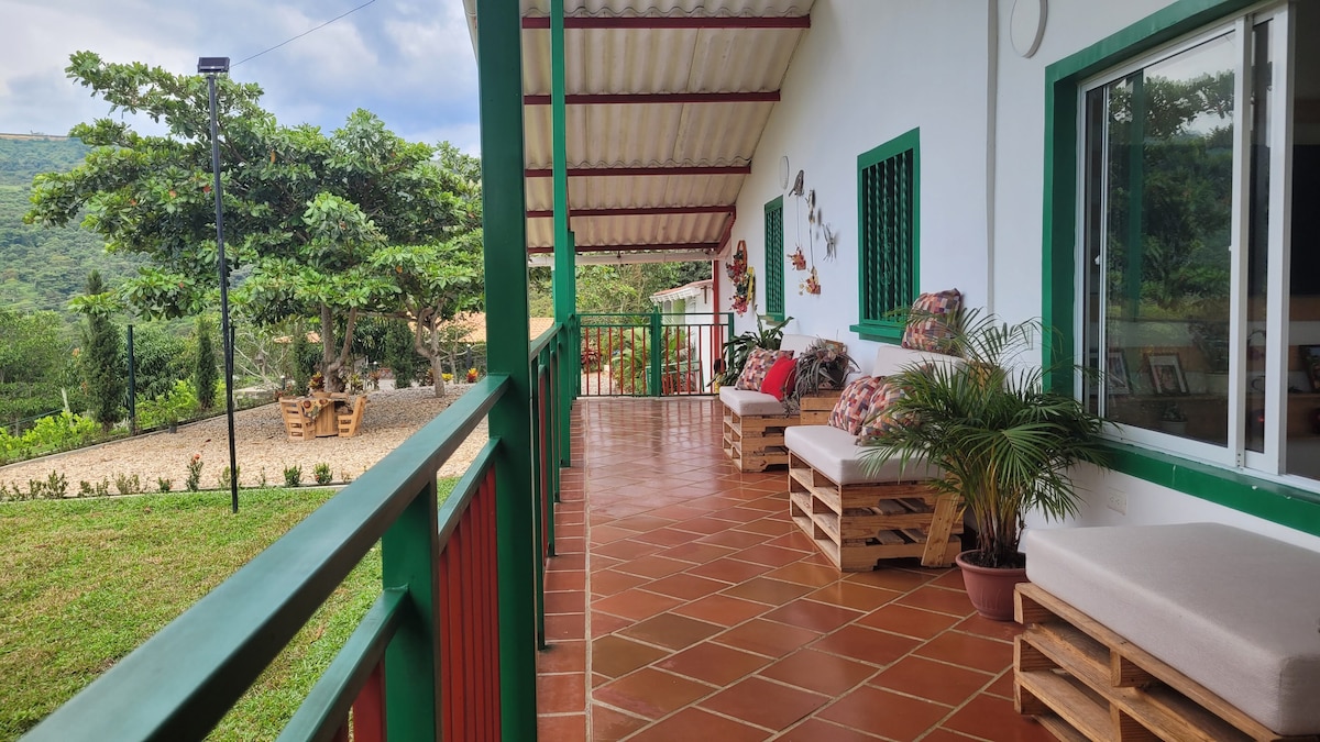 A covered porch is shown with seating made from wooden pallets, decorated with cushions. The tiled floor complements the vibrant green and red accents. Lush greenery and distant mountains are visible in the background, creating a serene outdoor experience.