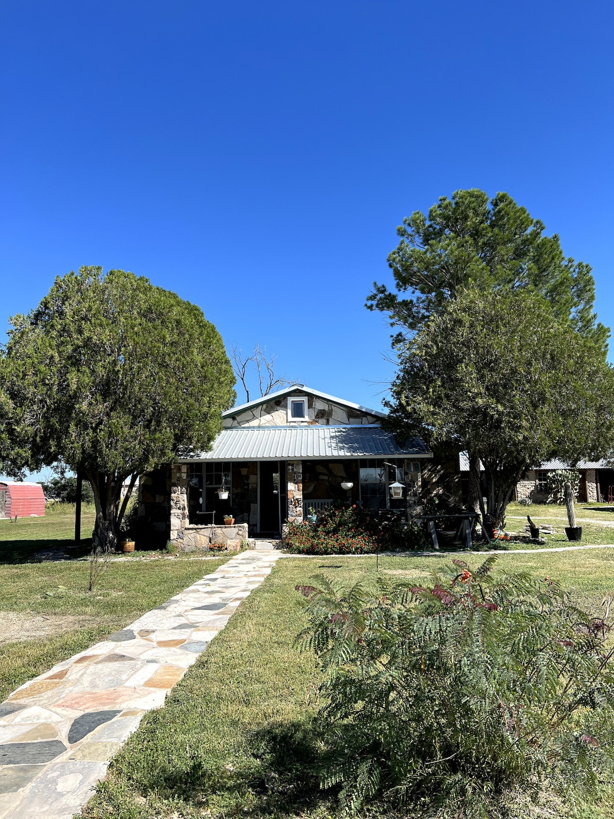 A charming house built from local rocks features a landscaped front yard with trees providing shade. A stone pathway leads to the entrance, flanked by vibrant greenery. The clear blue sky adds to the serene outdoor setting.