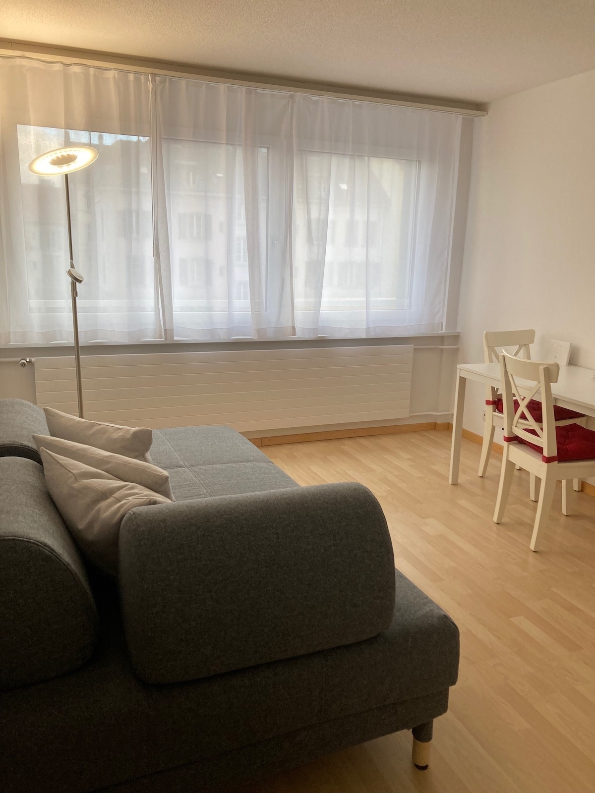 A well-lit living area features large windows with sheer white curtains, allowing natural light to fill the space. A cozy gray sofa with decorative pillows is positioned in the foreground, complemented by a compact white desk and a red chair in the background, enhancing the room's functionality.