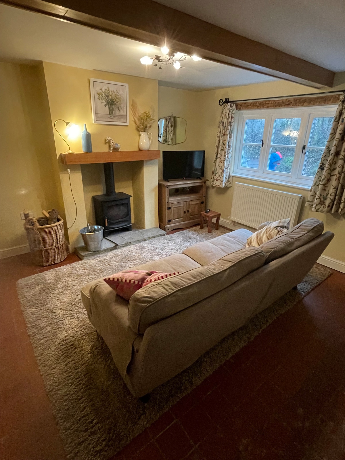 A warm lounge area features a beige sofa positioned on a soft rug, with a wooden coffee table nearby. A wood-burning stove is situated beneath a mantle, and a TV is placed against a wall. Natural light enters through a window, framed by patterned curtains.