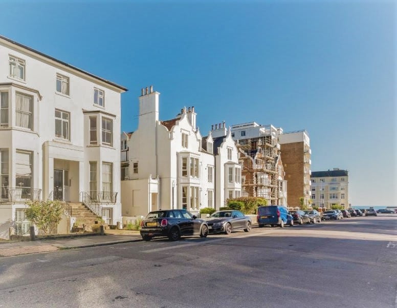 A row of white residential buildings is visible across the street, showcasing varying architectural styles. The street features parked cars, with the sea visible at the end of the road. Clear blue skies enhance the setting, contributing to a serene urban environment.