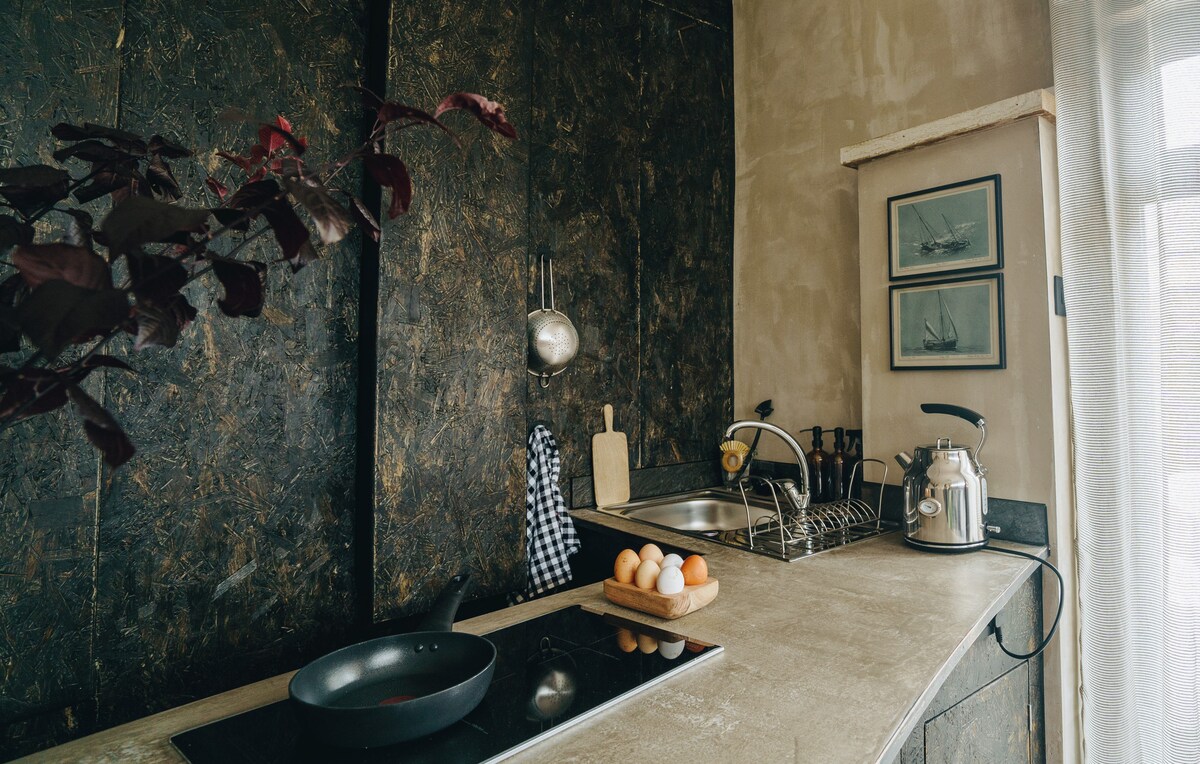 A well-equipped kitchen features sleek countertops and modern appliances. A frying pan sits on the stovetop, alongside a neatly arranged tray of eggs. Wall art depicting ships adorns the walls, complemented by a vase with dark leaves on the countertop.
