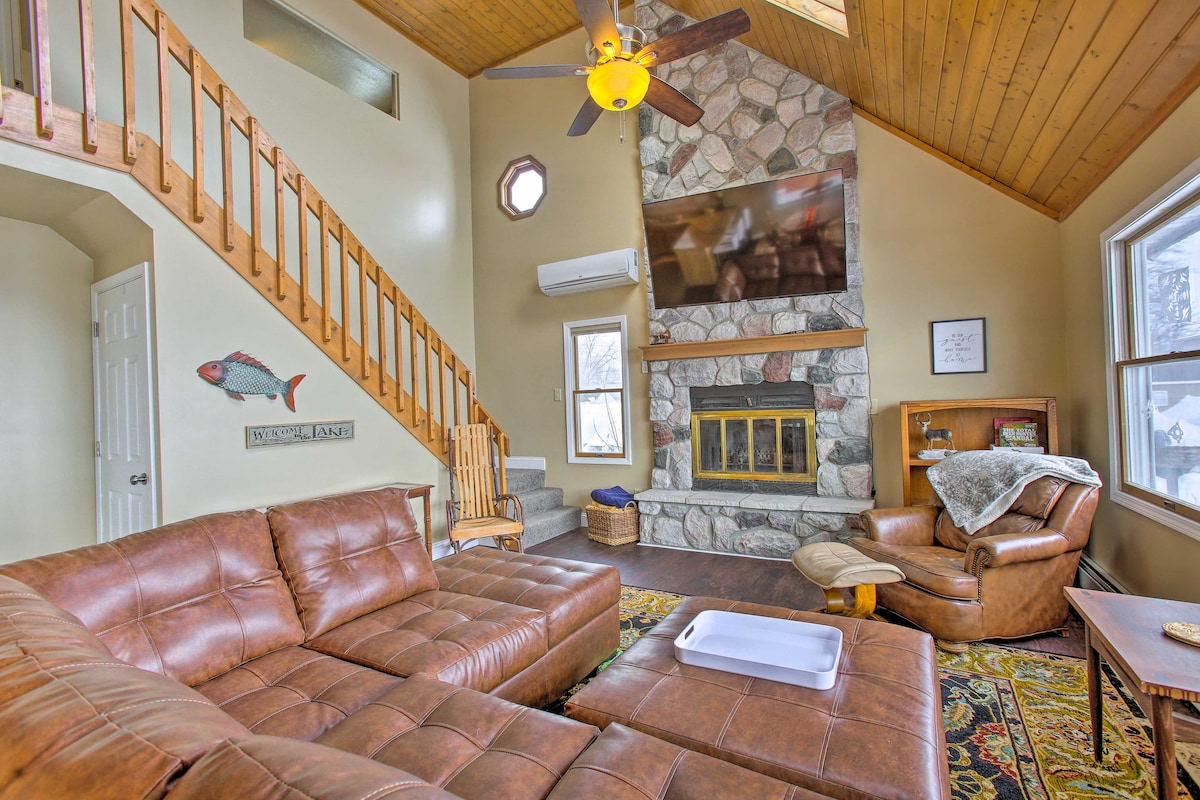 A spacious living area features a large leather sectional sofa, positioned to face a stone fireplace flanked by windows. A ceiling fan hangs above the area, while a staircase leads to an upper level. Natural light floods the space, enhancing the inviting atmosphere.