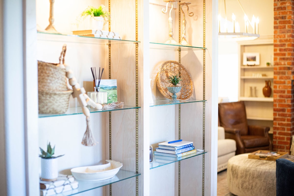 A decorative shelving unit is seen, featuring a variety of curated items such as woven baskets, small plants, and books. Soft lighting highlights the glass shelves, while the background displays a comfortable seating area with a leather chair and a cozy ottoman.