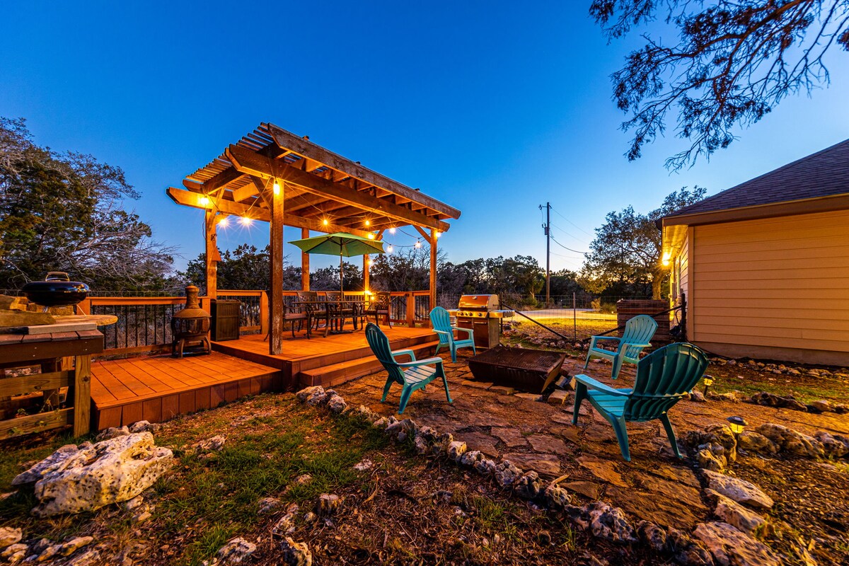 An outdoor gathering area is highlighted by a wooden pergola adorned with soft lighting. Comfortable seating is arranged around a stone firepit, with a spacious deck available for dining. Lush greenery surrounds the space, creating a peaceful atmosphere in the Texas Hill Country.