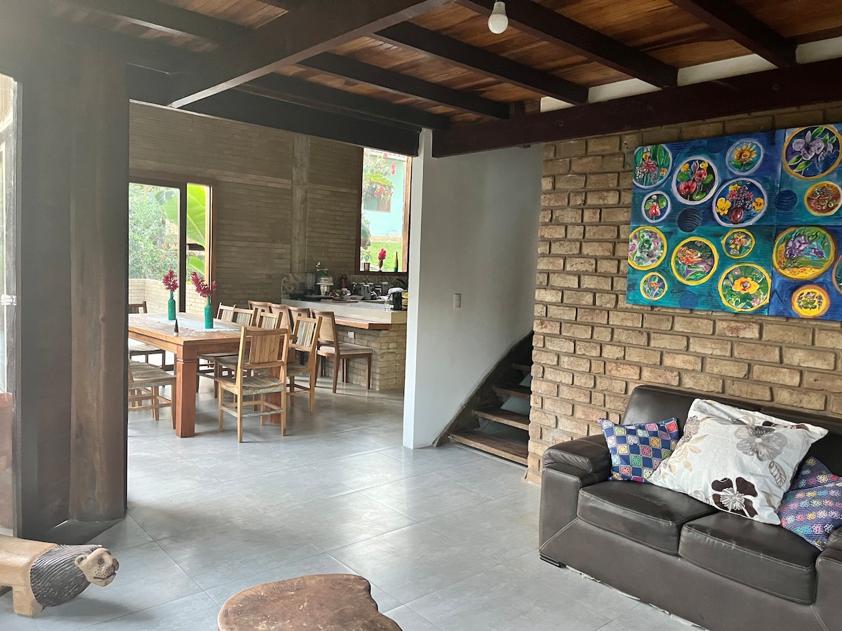 An open living space features a dark sofa with colorful cushions and a wooden table surrounded by several chairs. A staircase leads to an upper level, while large windows provide natural light and views of the surrounding greenery. A vibrant wall art piece adds a pop of color.