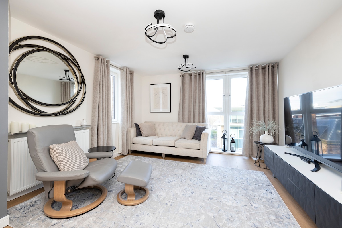 A modern living area is depicted, featuring a light-colored sofa and a matching ottoman. Large windows allow natural light to illuminate the space, with soft, neutral curtains adding warmth. A sleek entertainment unit holds a flat-screen TV, complemented by decorative elements and an abstract wall art piece.