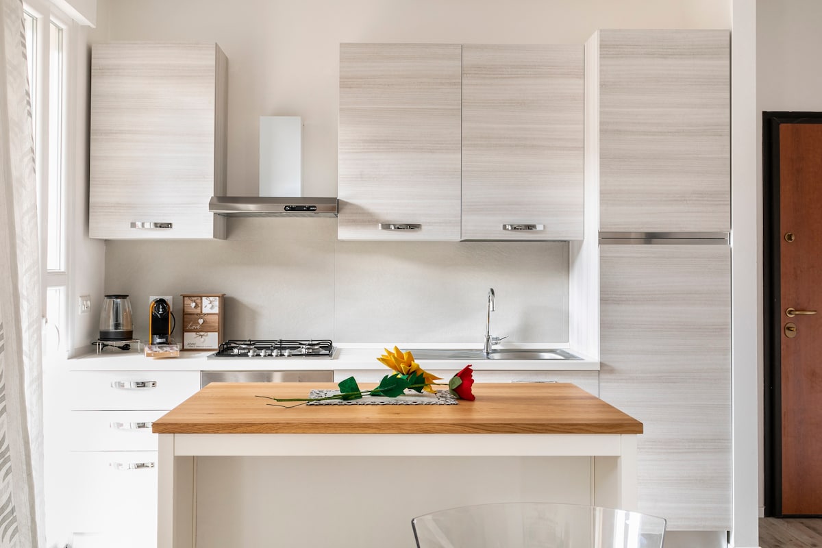 A modern kitchen is equipped with light wood cabinetry and stainless steel appliances. A central island with a wooden countertop features decorative flowers. Natural light streams through nearby windows, illuminating the clean and functional design.