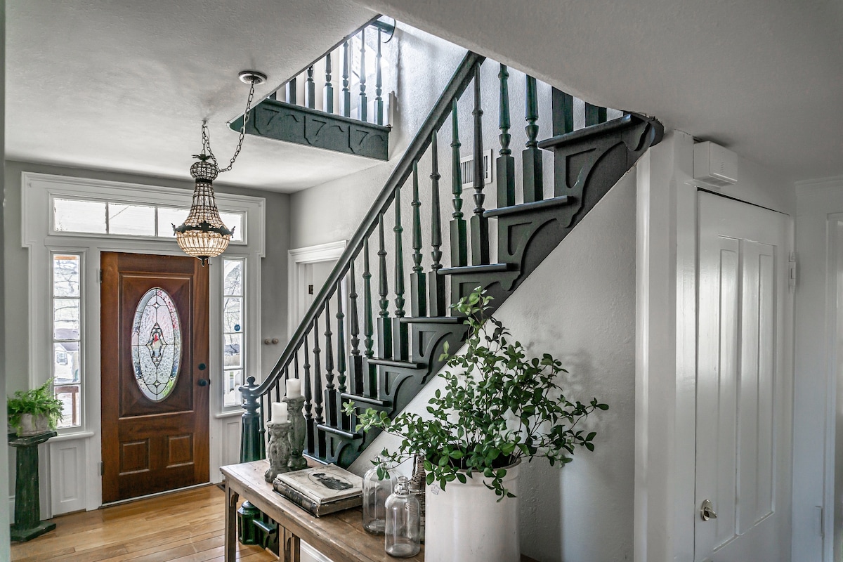 A welcoming entryway features a wooden door with stained glass detailing, offering natural light. The staircase, painted in a deep green, leads to the upper level. A wooden table adorned with decorative jars and a potted plant enhances the inviting atmosphere.