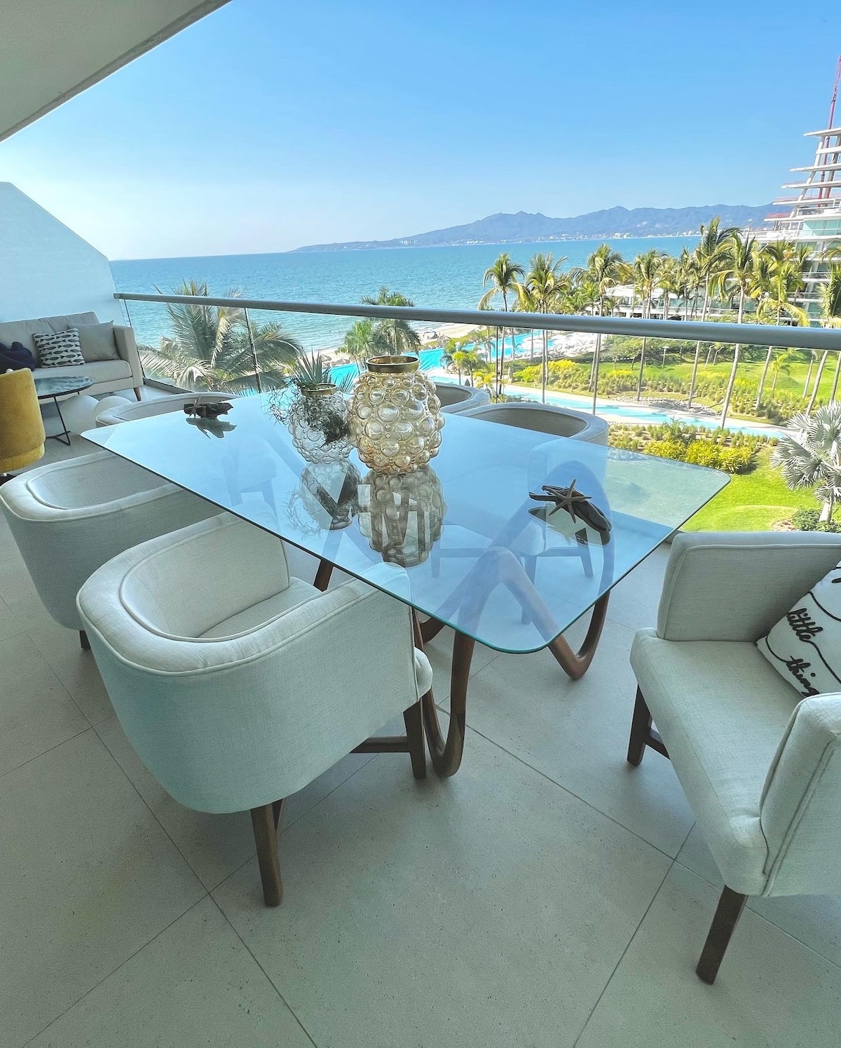 An outdoor dining area features a glass table surrounded by four upholstered chairs. A decorative vase sits at the center, while stunning ocean views and tropical scenery are visible in the background, showcasing the beauty of the beachfront location.