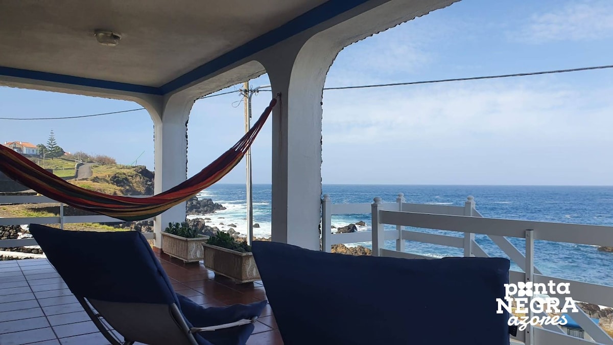 A covered terrace is seen overlooking the ocean, with a hammock hung for relaxation. Two lounge chairs are positioned on the tiled floor. The coastline and blue sea are visible in the background, providing a scenic and serene view.