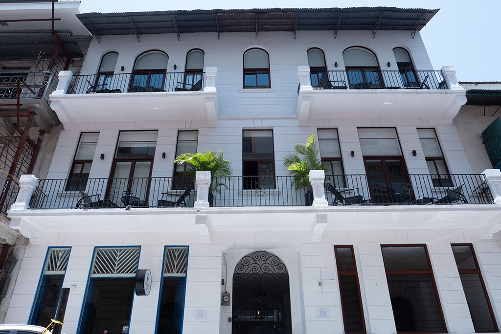 A colonial-style building showcases a white facade with arched windows and balconies adorned with plants. The entrance is framed by decorative columns and features black railings. The structure reflects a blend of historical architecture and modern design elements.