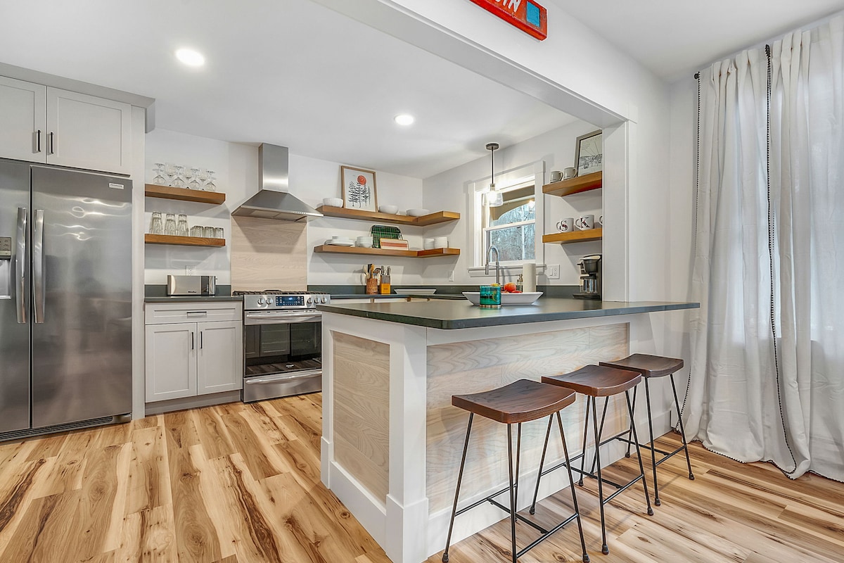 A modern kitchen features stainless steel appliances and sleek cabinetry, accented by open shelving displaying dishware. A spacious island with seating for three is complemented by hardwood flooring. Natural light enters through a window, enhancing the inviting atmosphere.