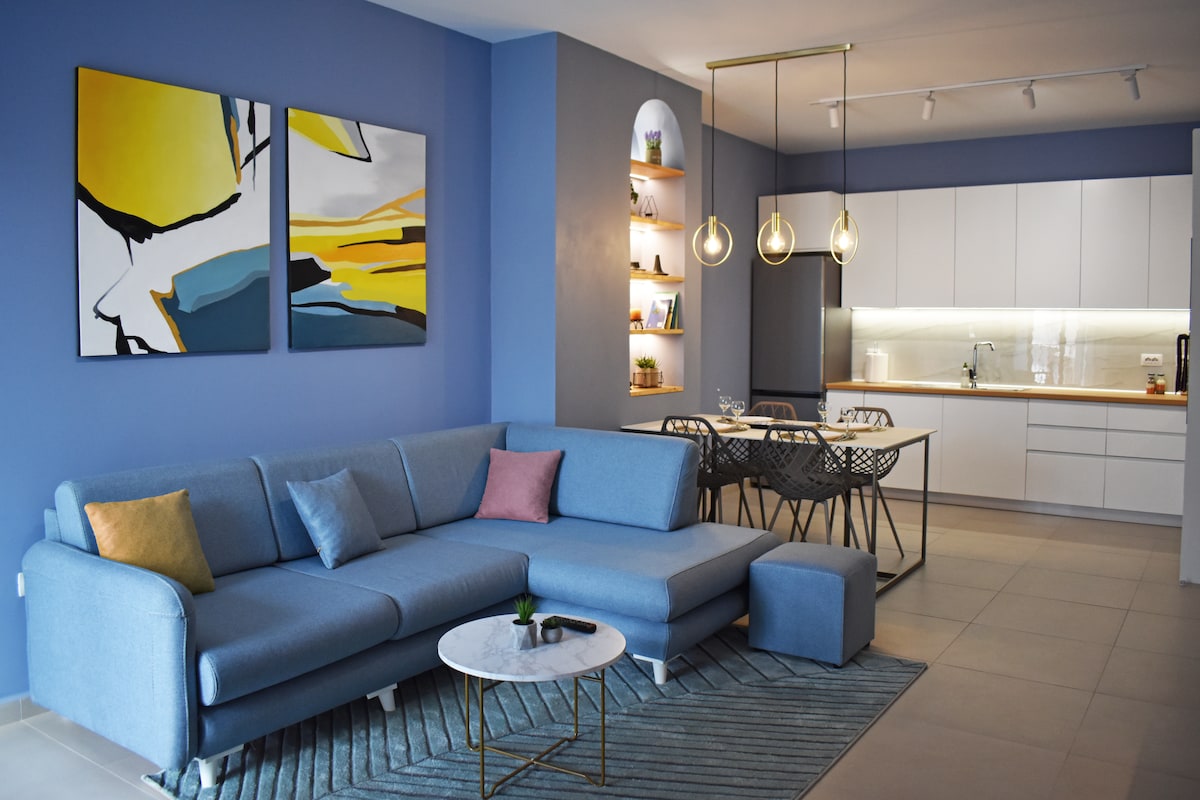 A modern living area features a blue sectional sofa with decorative pillows and a marble coffee table. In the background, a sleek kitchen showcases white cabinetry, illuminated shelving, and contemporary pendant lighting above a dining table with stylish chairs. Bright artwork adorns the wall.