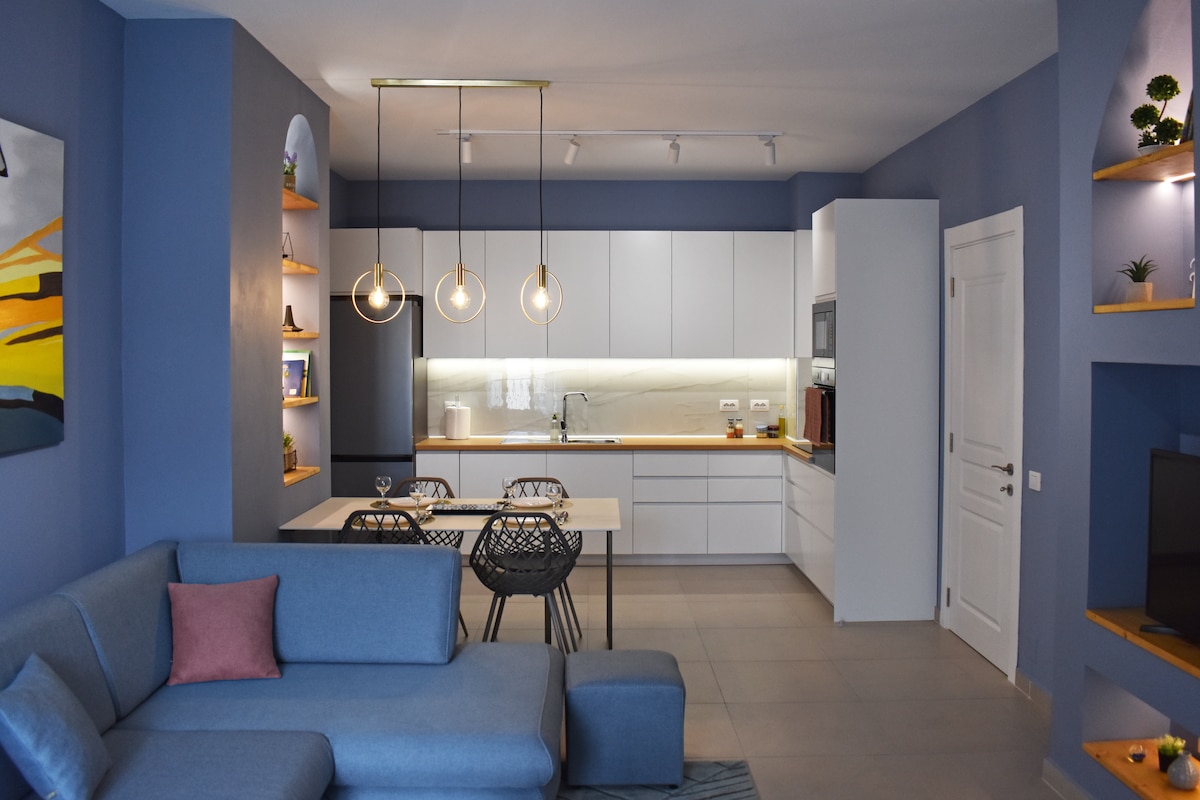 A modern kitchen area features sleek cabinetry and a well-lit countertop. Three pendant lights hang above a dining table with four chairs. A comfortable sectional sofa is positioned in the adjacent living area, complemented by a decorative pillow.