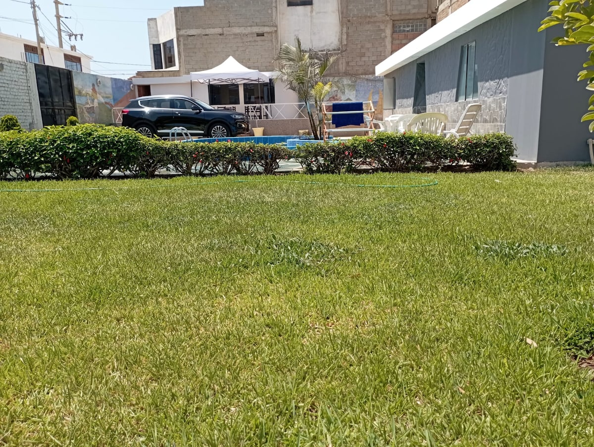 A well-maintained garden features lush green grass and neatly trimmed hedges. In the background, a swimming pool and outdoor seating area are visible, along with a white tent providing shade. A vehicle is parked nearby, enhancing accessibility.
