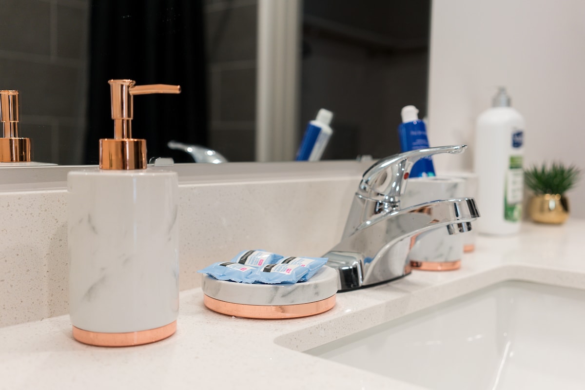 A clean bathroom countertop is presented, featuring a sleek, modern faucet alongside marble and rose gold accessories. Soap and lotion dispensers coordinate with a small tray holding packaged toiletries. Soft lighting illuminates the serene space, enhancing the contemporary style.