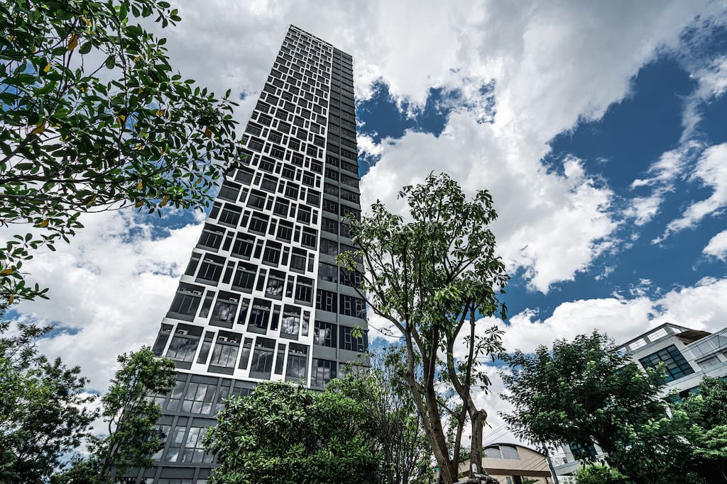 A tall, modern building reaches toward the sky, surrounded by lush greenery. The structure's facade features large, geometric windows reflecting the clouds above, creating a striking visual against the vibrant blue sky.