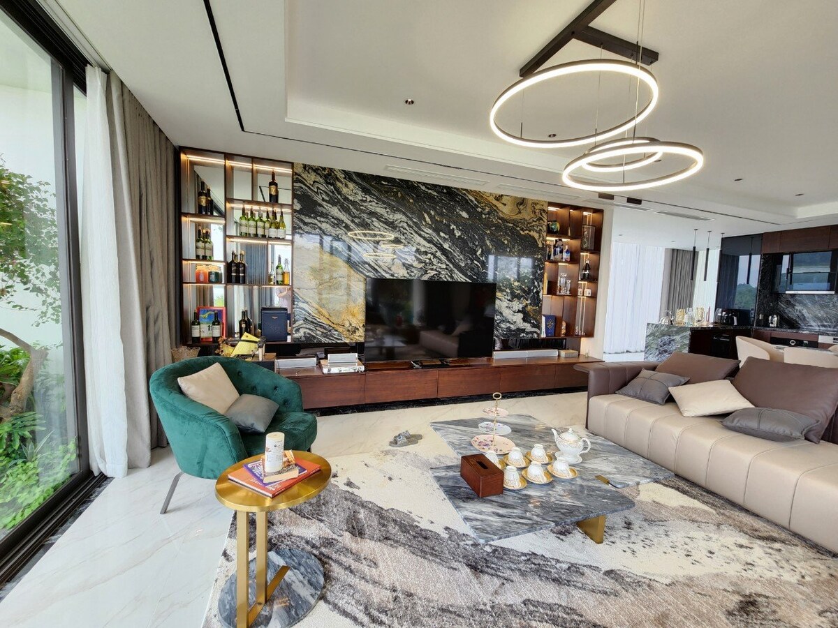 The living area features a contemporary design with a large wall-mounted television, framed by elegant cabinetry containing various bottles. A plush sectional sofa faces a decorative coffee table, which is surrounded by a soft, patterned rug. Natural light floods the space through expansive glass doors.