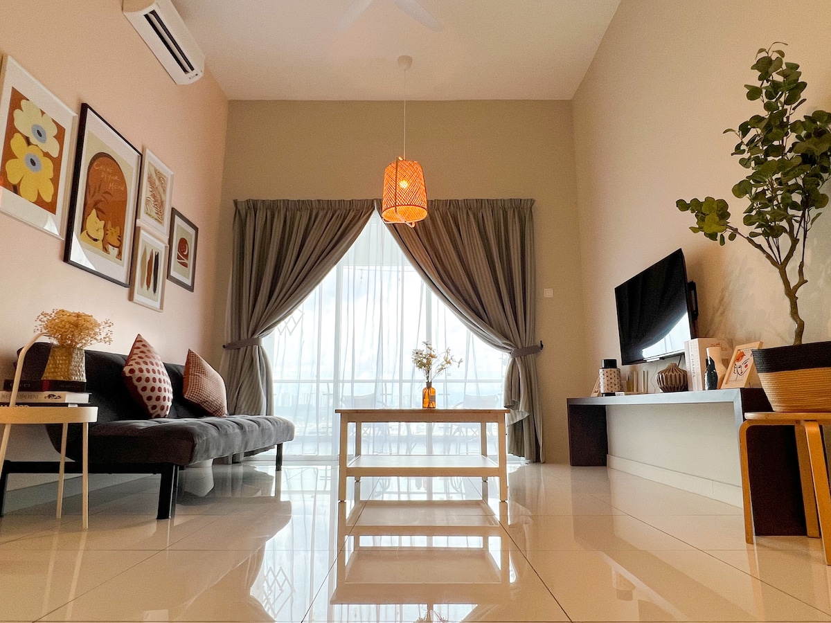 A living area features a dark sofa and a white coffee table placed centrally. Large windows allow natural light to brighten the space, adorned with soft curtains. Decorative elements include framed artwork on the walls and a small plant beside the television unit.