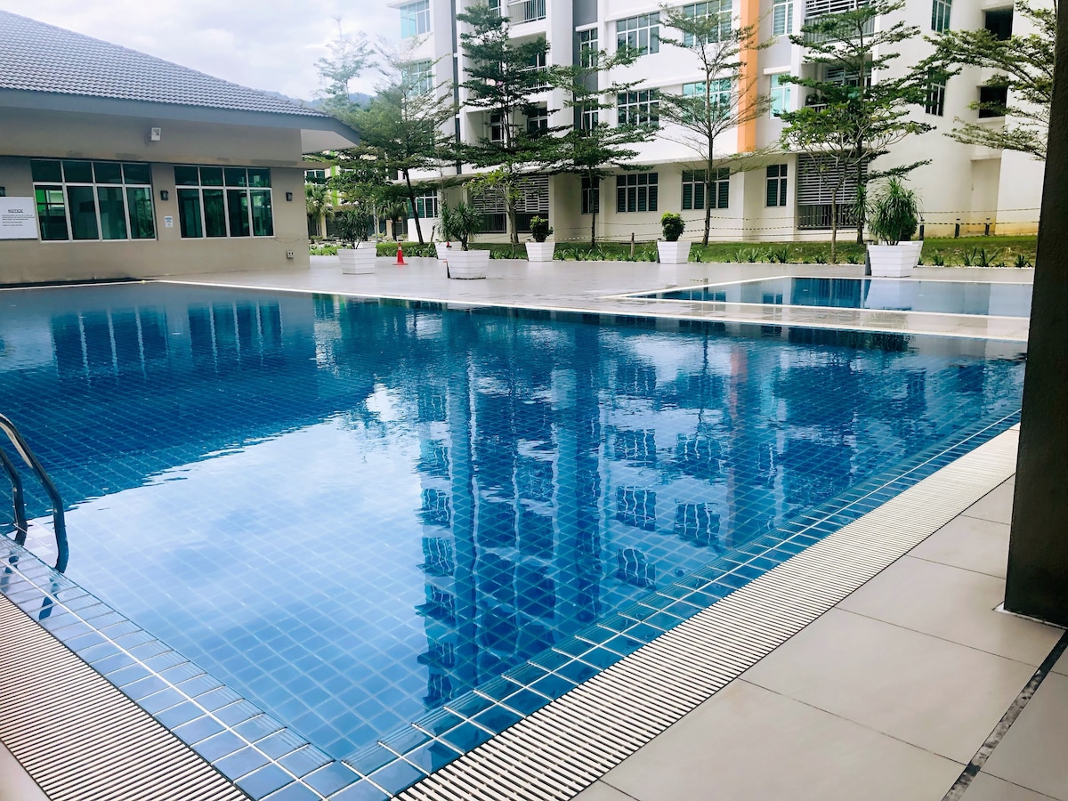 A large swimming pool features crystal-clear water reflecting the surrounding buildings. Greeneries and white planters line the edge, adding a touch of nature to the area. Sunlight creates a tranquil ambiance around the spacious pool deck.
