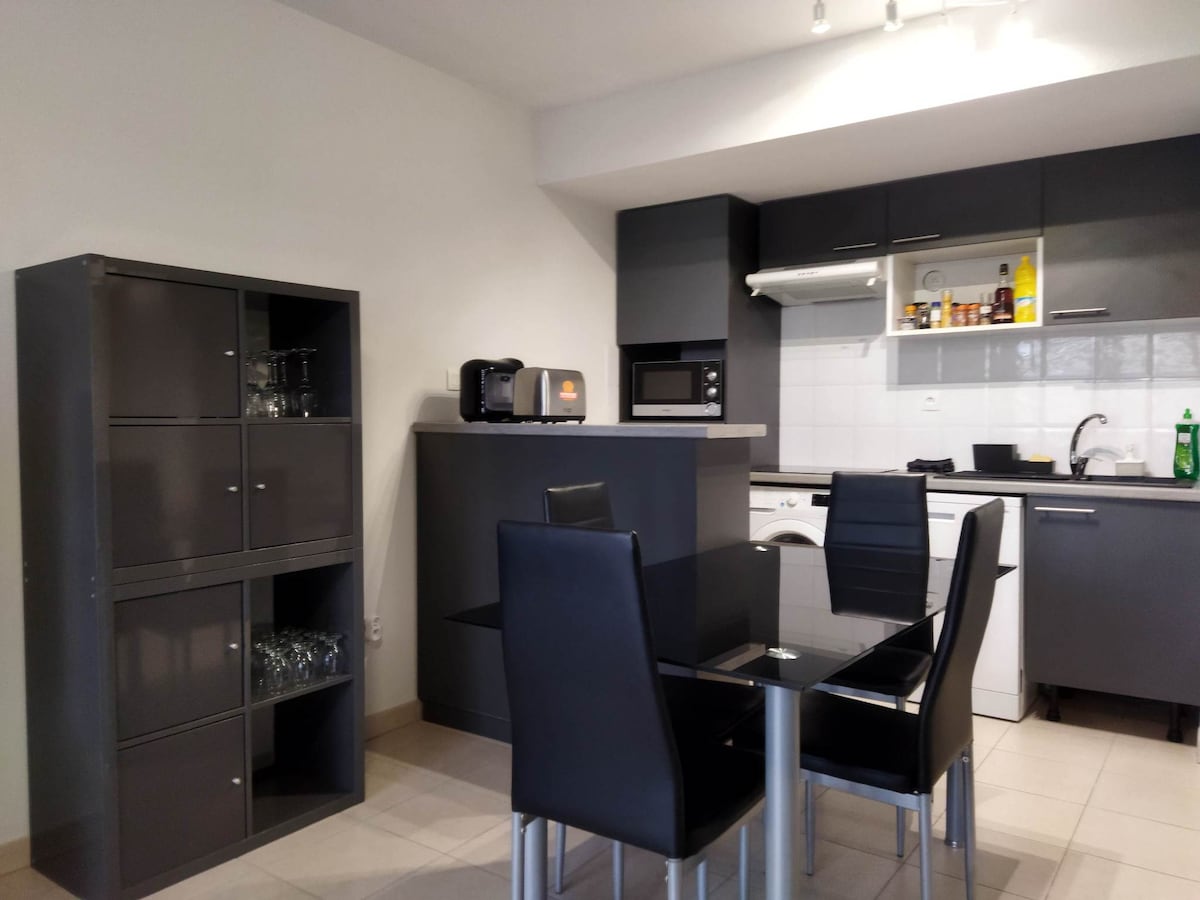 A modern kitchen area is equipped with sleek dark cabinetry and stainless steel appliances. A glass dining table is surrounded by four black chairs. Kitchen essentials are visible, including a microwave, coffee maker, and an array of cooking supplies in the organized space.