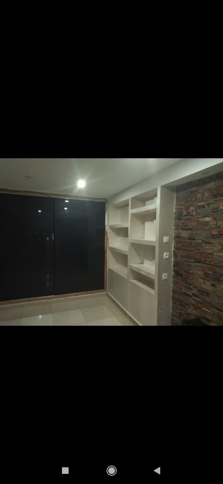 A modern living space is highlighted by a textured stone wall and a large glass door allowing natural light. Built-in shelves are integrated into one wall, providing functional storage. The floor features glossy tiles that add to the room's contemporary aesthetic.