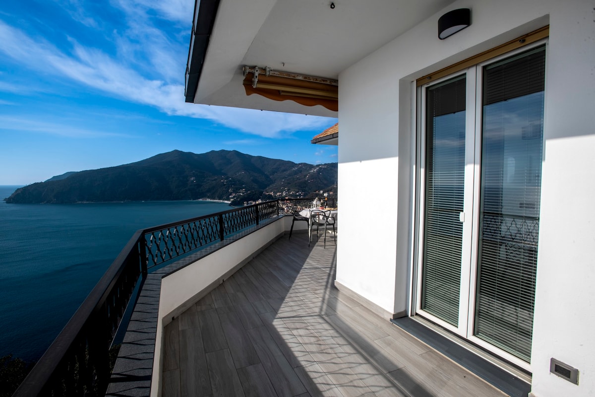 A spacious terrace is seen overlooking the bay, with panoramic views of the surrounding mountains and sea. Large glass doors connect the interior space to the terrace, allowing for natural light to fill the area. Outdoor seating is arranged for enjoying meals al fresco.