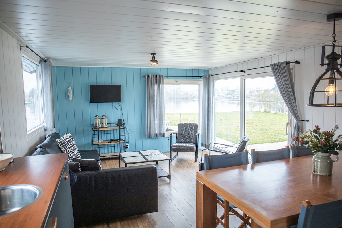 A spacious living area is showcased, featuring a large corner sofa, two armchairs, and a coffee table. Large windows provide views of the outdoor landscape, allowing natural light to fill the room. A dining table for six is also visible, enhancing the communal space.