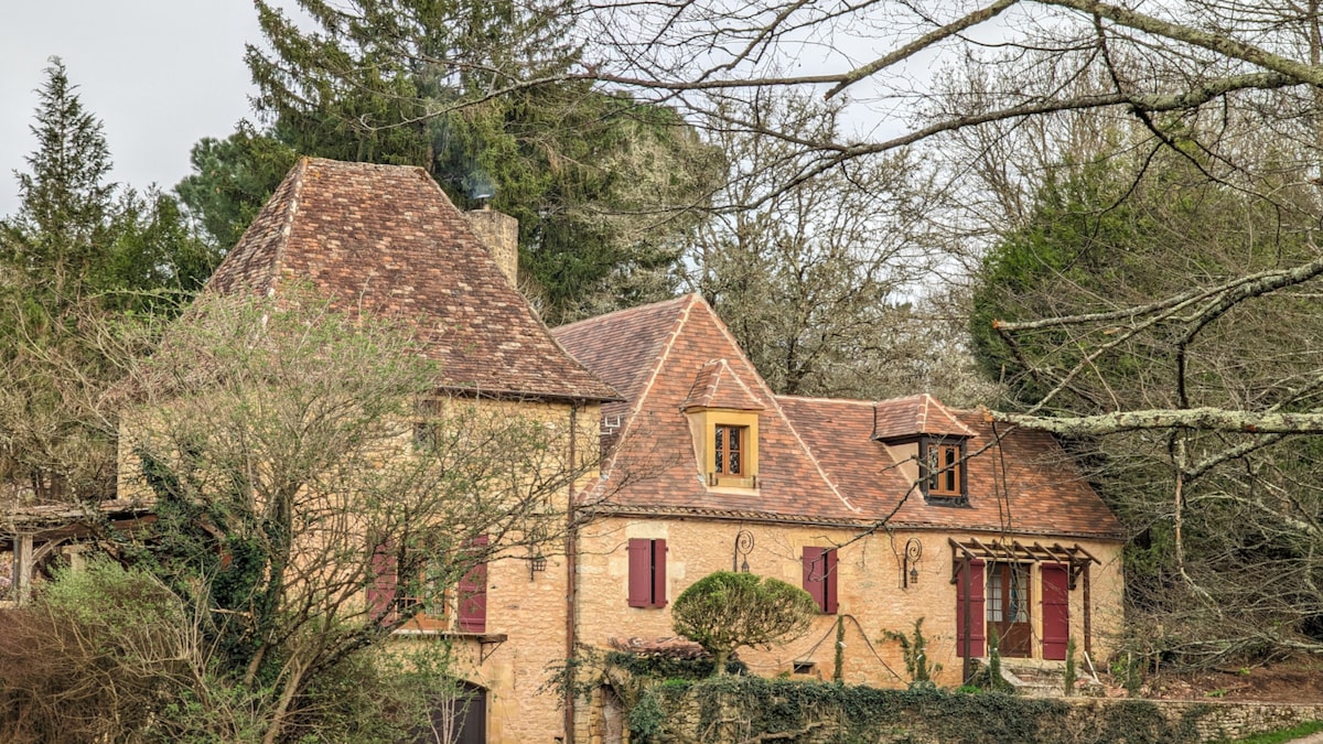 The historic stone residence features a distinctive tiled roof and red shutters. Surrounding greenery includes trees and shrubs, creating a natural backdrop. Architectural details showcase traditional elements, enhancing the charm of the home within its tranquil setting.